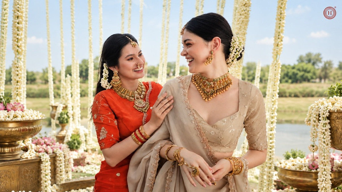 Two women in traditional Indian attire posing outdoors. One wears a vibrant red saree with gold embroidery and gold jewelry, while the other wears a beige saree with gold embroidery and matching gold jewelry.