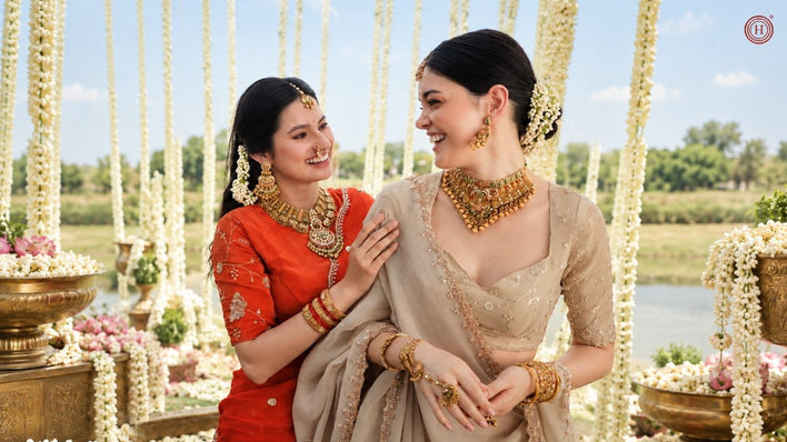 Two women in traditional Indian attire posing outdoors. One wears a vibrant red saree with gold embroidery and gold jewelry, while the other wears a beige saree with gold embroidery and matching gold jewelry.